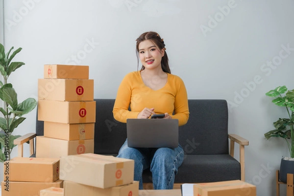 Fototapeta E-commerce Hustle: Young Asian entrepreneur managing orders and logistics on phone and tablet, surrounded by shipping boxes in her home office.	