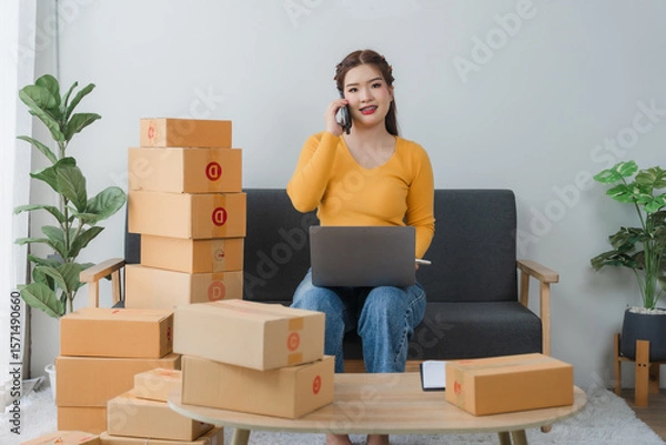 Fototapeta E-commerce Hustle: Young Asian entrepreneur managing orders and logistics on phone and tablet, surrounded by shipping boxes in her home office.	