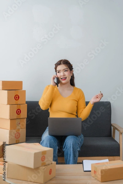 Fototapeta E-commerce Hustle: Young Asian entrepreneur managing orders and logistics on phone and tablet, surrounded by shipping boxes in her home office.	
