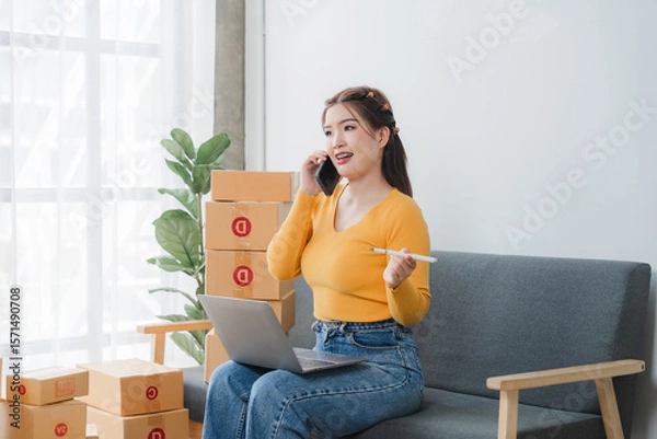 Fototapeta E-commerce Hustle: Young Asian entrepreneur managing orders and logistics on phone and tablet, surrounded by shipping boxes in her home office.	