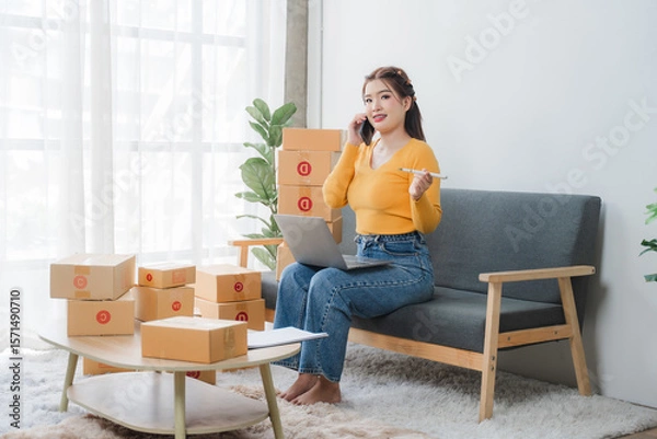 Fototapeta E-commerce Hustle: Young Asian entrepreneur managing orders and logistics on phone and tablet, surrounded by shipping boxes in her home office.	