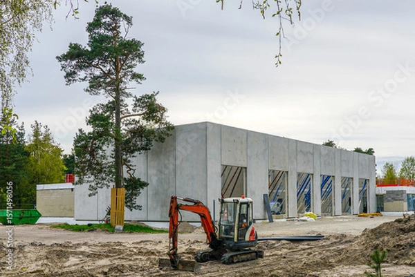 Fototapeta Concrete Commercial Building Under Construction with Excavator and Protected Tree on Site