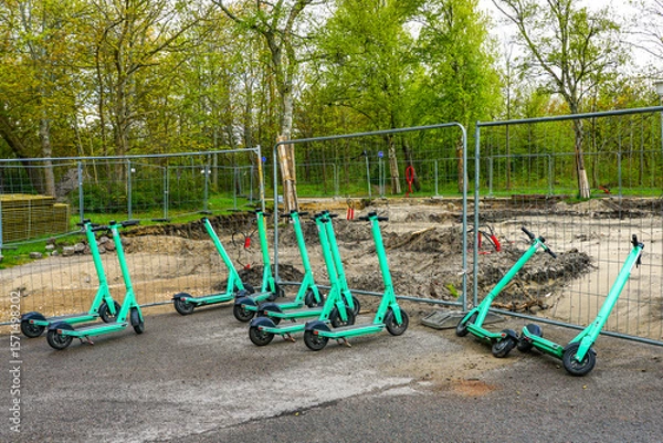 Fototapeta Shared Electric Scooters Parked at a Construction Site with Safety Fence and Tree Background