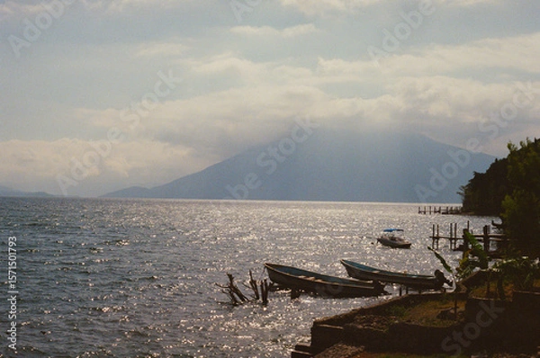 Fototapeta Scenic view of Lake Atitlán with still waters and a distant volcano. Boats are docked along the shoreline, highlighting a serene and peaceful natural landscape during a sunny day. Taken on 35mm Film.