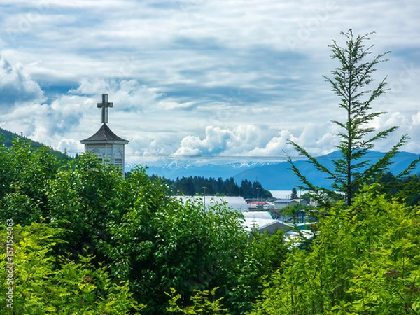 Obraz A cross atop the steeple of a hillside church in the port town of Wrangell, Alaska, with a view of distant mountains, some capped with snow, on a cloudy morning early in summer