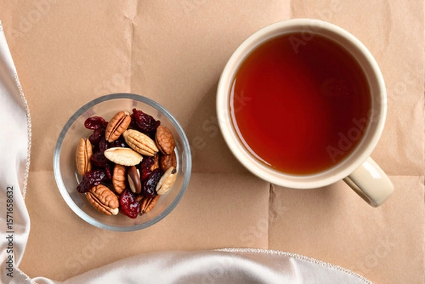 Fototapeta cozy top-view scene with a cup of black tea, a bowl of mixed nuts and dried cranberries, and a pink silk scarf arranged on a brown craft paper background for a relaxing break