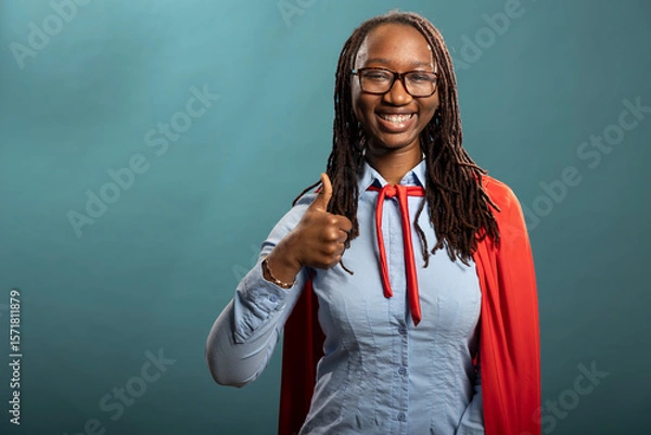 Fototapeta Cheerful black heroine giving a thumbs up, portraying positivity and agreement. African american woman in red cape showing approval gesture and looking at camera with bright smile.
