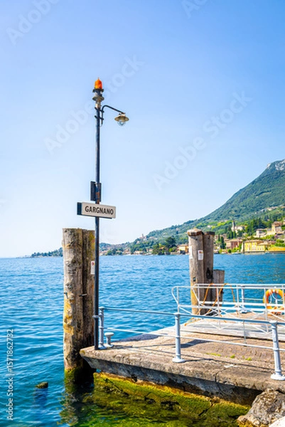 Fototapeta Streetlamp and Gargnano signpost by Lake Garda, Italy, with clear blue sky and calm lake waters, ideal for travel and destination visuals