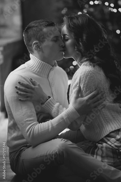 Fototapeta Young couple staying at home together on holiday evening and sitting on floor in front of christmas tree and hugging. Cute romantic lovers in white pullovers