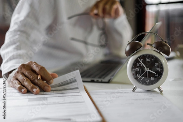 Fototapeta selective focus on alarm clock, asian businessman review business document using laptop working on table before work time end in a day,  time management concept, routine work, office hour