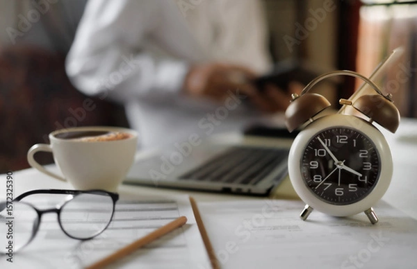 Fototapeta select focus on alarm clock on table near laptop, documents and eyeglasses and blur businessman working behind, time management concept, routine work, office hour