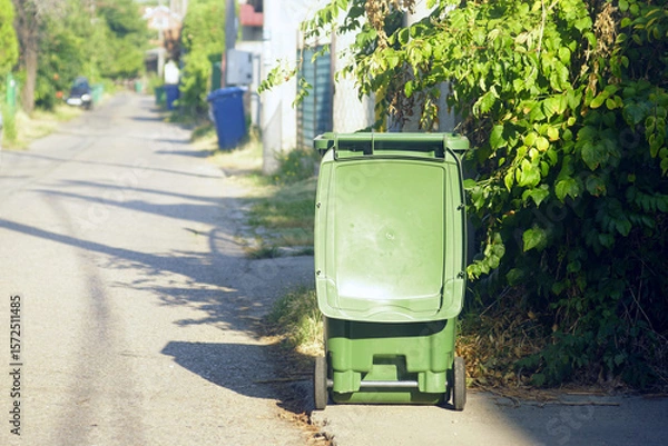 Fototapeta Waste collection and sorting in the everyday life of a small European city: a green wheeled trash bin with an open lid on a deserted sunny street and blue garbage containers in the distance.