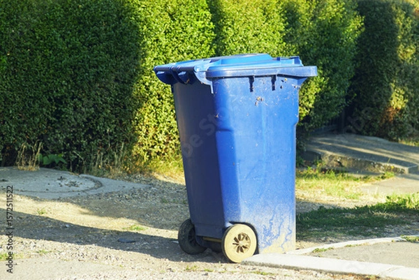 Fototapeta Blue garbage container with lid and wheels on a residential street in summer. Symbolizing of modern recycling and sustainability awareness. Waste management in suburban or small-town neighborhoods