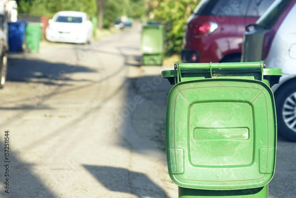 Fototapeta Green street trash bin with open lid standing on the side of the road next to cars, close-up photo. Concept of how small towns handle growing ecological challenges through daily waste disposal