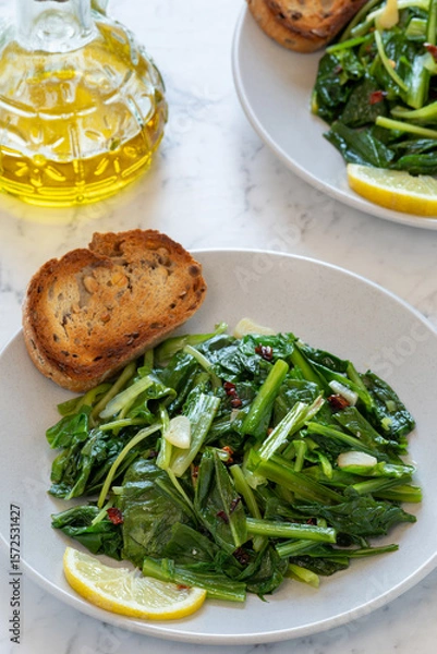 Obraz Cooked chicory greens sautéed in olive oil, garlic, and hot pepper flakes - a traditional Italian side dish served on a plate with toasted bread and a slice lemon. A bottle of olive oil on the side.