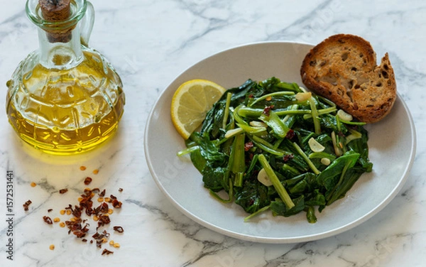 Obraz Sauteed Chicory Greens - a traditional Italian side dish on a plate served with toasted bread and a slice of lemon. A bottle of olive oil and some scattered hot pepper flakes on the side.