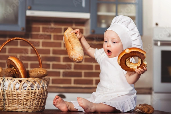 Fototapeta Funny little cook in kitchen with bakery. Basket with loafs a bread and bagels