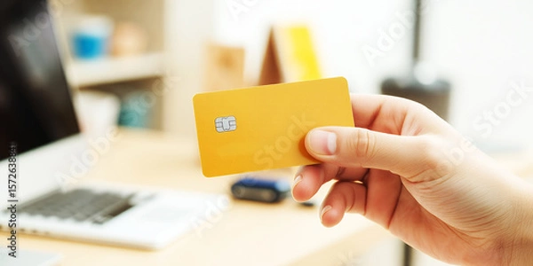 Obraz A hand holding a yellow credit card in front of a laptop on a desk in a bright room on transparent background