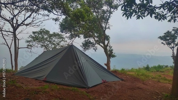 Obraz A dark-colored flysheet tent stands on a misty mountain peak, surrounded by tall trees and foggy skies, creating a serene and secluded camping atmosphere.