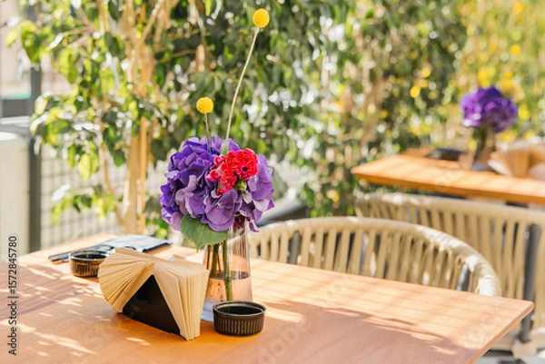 Fototapeta Glass vase with purple hydrangeas, red carnations, and yellow billy balls on an outdoor cafe table in bright sunlight.
