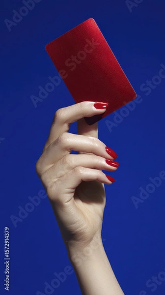Obraz Elegant Hand Presenting a Shiny Red Card Against a Vivid Blue Background with Glamorous Nail Polish