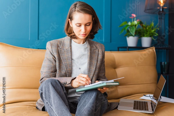 Fototapeta Beautiful brunette sitting on the sofa and taking notes while working on her laptop.