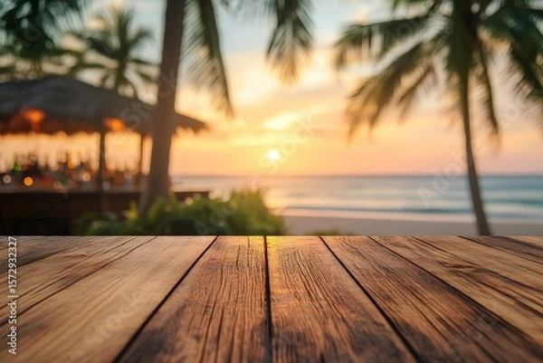 Fototapeta Wooden table surface with a blurred background of a tropical beach bar at sunset.