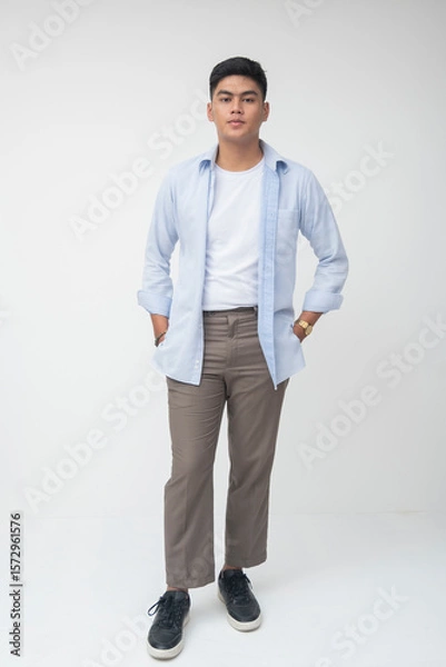 Fototapeta Southeast Asian Gen Z man stands casually with hands in pockets, wearing light blue long sleeves, gray pants, and minimalist shoes with a calm, neutral expression.
