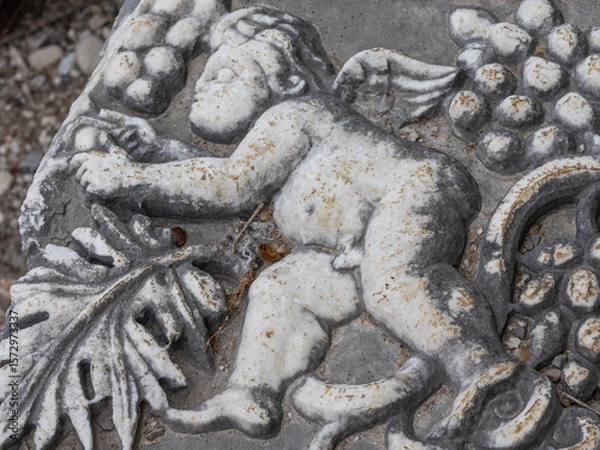 Fototapeta Bas-relief on the surface of a stone block in the ancient city of Perge in Turkey. An image of an angel and grapes on a stone surface. Grape leaves and berries next to an angel on a stone.