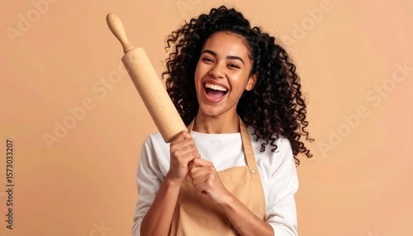 Fototapeta Cheerful Woman in Apron Holds Rolling Pin in Studio Setting Against Tan Background