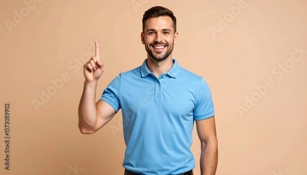 Fototapeta Smiling Man in Blue Polo Shirt Pointing Upward with Neutral Background in Studio Light