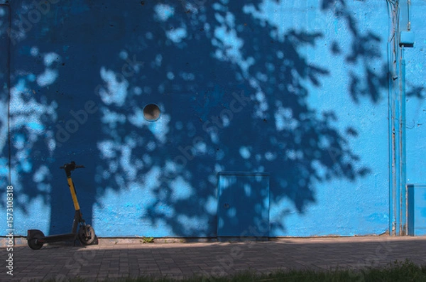 Fototapeta a blue wall of a building with a shadow from a tree and an electric scooter nearby
