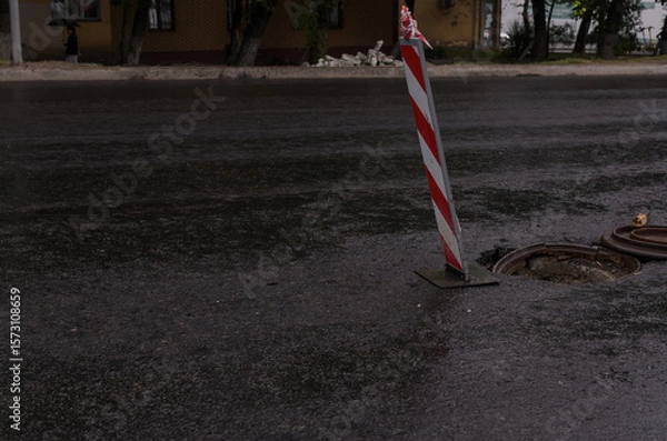 Fototapeta red and white road fence post in front of an open road hatch on a rainy evening