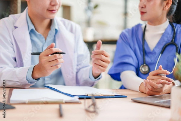 Fototapeta Two healthcare professionals, one in white coat and one in blue scrubs, discuss patient information at desk with documents and laptop, showing teamwork and focus in medical office