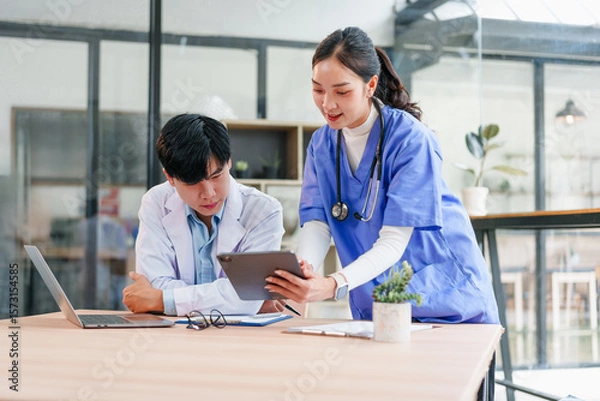 Fototapeta Two healthcare professionals, one male and one female, collaborate in modern office setting, reviewing patient information on digital tablet, displaying teamwork and focus
