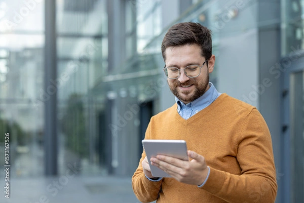 Fototapeta Close-up photo of a young smiling man standing outside and using a tablet in his hands