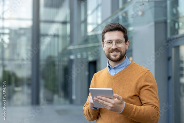Fototapeta Portrait of a young businessman wearing glasses standing outside an office center, holding a tablet in his hands and looking at the camera with a smile