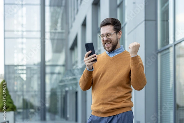 Fototapeta Happy young male businessman standing near the building, looking at the phone screen and rejoicing in success