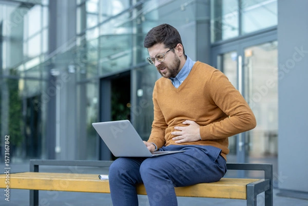 Fototapeta A young businessman, twisted in pain, sits on a bench outside near a building, working on a laptop, holding his stomach with his hand