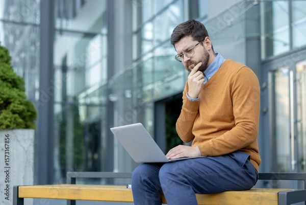 Fototapeta A thoughtful young man sits on a bench on the street near an office building and looks intently at the laptop screen