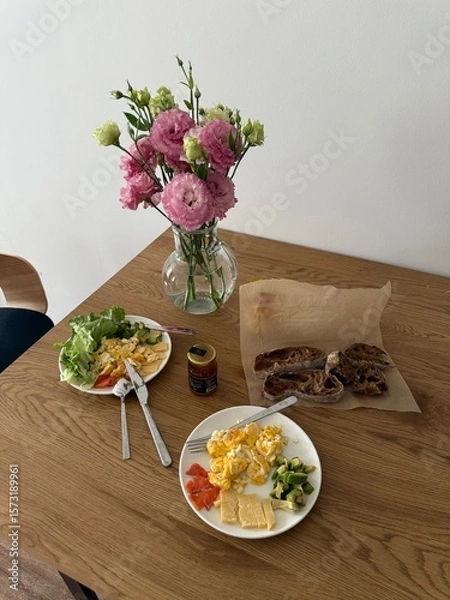 Fototapeta Two breakfast plates with eggs, cheese, avocado, and vegetables on a wooden table. Sliced bread and a vase with flowers are also visible.