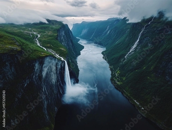 Fototapeta A dramatic aerial scene of a deep Norwegian fjord framed by towering cliffs and lush greenery, with a powerful waterfall cascading into a serene, dark blue river below.

