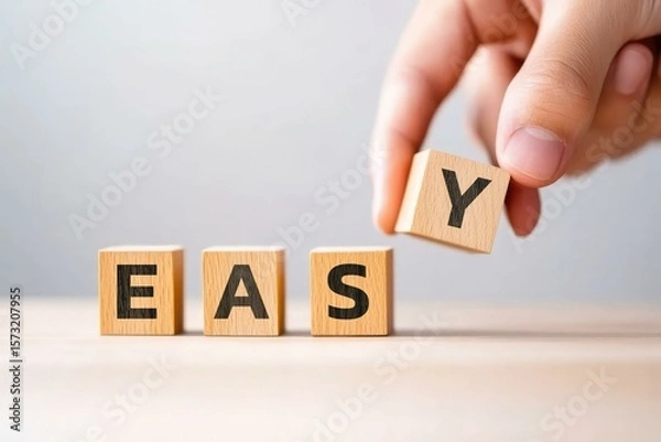 Fototapeta A close-up image of a hand placing the letter 'Y' onto wooden blocks spelling 'EASY', illustrating simplicity and ease in learning or tasks, perfect for educational or motivational materials.