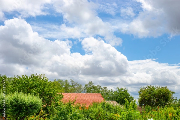 Fototapeta Countryside landscape. view of a house with a red tile roof surrounded by lush green trees under a cloudy blue sky.