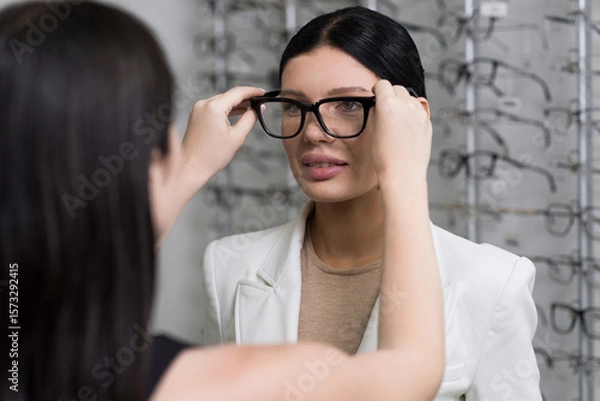 Fototapeta Attractive Caucasian young brunette woman in white suit trying on and choosing eyeglasses for buying at an optical store. Lifestyle, optometry, eyesight, assortment of glasses