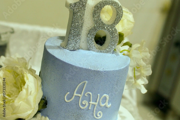 Fototapeta A two-tiered blue and white birthday cake decorated with a pearl ribbon and white and yellow flowers with the female name Ana written in Cyrillic script