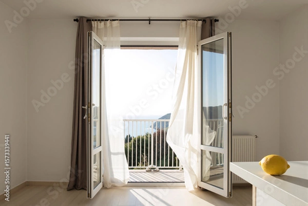 Fototapeta Open balcony door with a white curtain blowing in a gentle breeze, creating a bright, airy, and peaceful atmosphere in a minimalist room with a single fresh lemon on a table