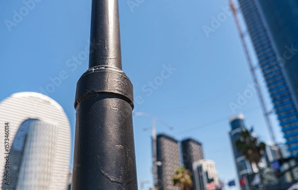 Fototapeta A minimalist city scene captured with foreground focus on a black metal pole against blurred modern skyscrapers and a clear blue sky. Perfect for design projects exploring urban life. Selective focus.