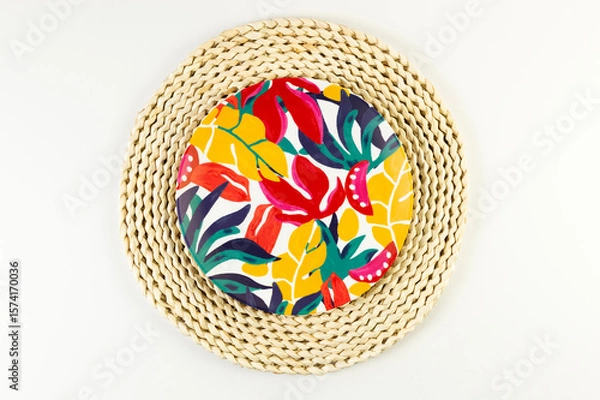 Fototapeta Top view of brown wicker table mat, colorful plate on white background. Straw round tray, exotic plant plate, summer picnic. Flat lay, copy space. 