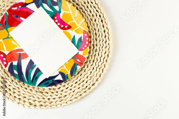 Fototapeta Top view of brown wicker table mat, white card, colorful plate on white background. Straw round tray, exotic plant plate, summer picnic. Flat lay, copy space. 